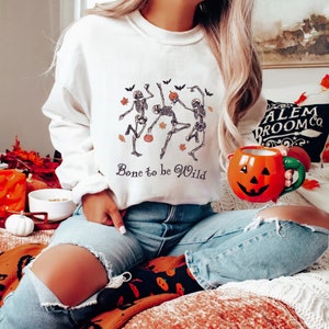 K&ouml;nnte beinhalten: Wei&szlig;er Pullover mit einem Motiv von drei Skeletten, die mit K&uuml;rbissen und Flederm&auml;usen tanzen. Der Text "Bone to be Wild" ist unter dem Motiv gedruckt.