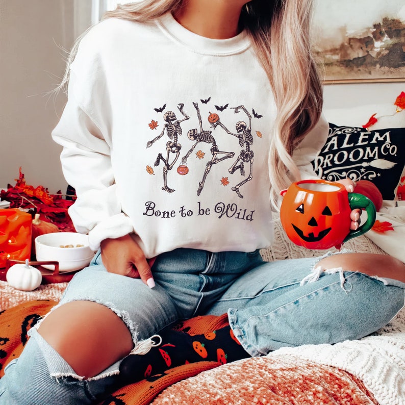 K&ouml;nnte beinhalten: Wei&szlig;er Pullover mit einem Motiv von drei Skeletten, die mit K&uuml;rbissen und Flederm&auml;usen tanzen. Der Text "Bone to be Wild" ist unter dem Motiv gedruckt.