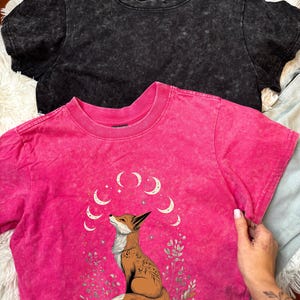 May include: Two short-sleeved t-shirts. One is black, the other is hot pink with a fox graphic and crescent moon design. The fox is brown and white, surrounded by floral elements. The shirts have a textured, washed appearance.