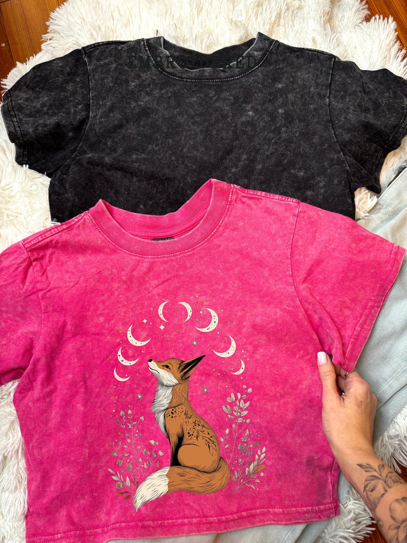 May include: Two short-sleeved t-shirts. One is black, the other is hot pink with a fox graphic and crescent moon design. The fox is brown and white, surrounded by floral elements. The shirts have a textured, washed appearance.
