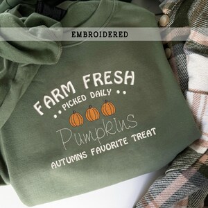 Op de afbeelding: Een olijfgroene sweatshirt met bovenaan het woord "EMBROIDERED". De sweatshirt heeft de tekst "FARM FRESH PICKED DAILY" en het woord "Pumpkins" met drie oranje pompoenillustraties. De zin "AUTUMNS FAVORITE TREAT" staat onderaan.