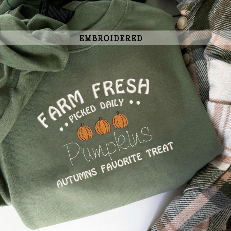 Op de afbeelding: Een olijfgroene sweatshirt met bovenaan het woord "EMBROIDERED". De sweatshirt heeft de tekst "FARM FRESH PICKED DAILY" en het woord "Pumpkins" met drie oranje pompoenillustraties. De zin "AUTUMNS FAVORITE TREAT" staat onderaan.