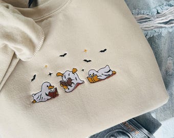 Puede incluir: Sudadera beige con diseño bordado de Halloween. Presenta tres patos fantasmas leyendo libros, con murciélagos y estrellas arriba. La sudadera está doblada, con un vistazo a unos vaqueros y un accesorio con temática de calaveras.