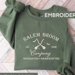 K&ouml;nnte beinhalten: Gr&uuml;ner Pullover mit dem Text "Salem Broom Company est. 1626 Enchanted + Handcrafted" und einer Grafik von zwei gekreuzten Besen mit Flederm&auml;usen, die um sie herum fliegen.