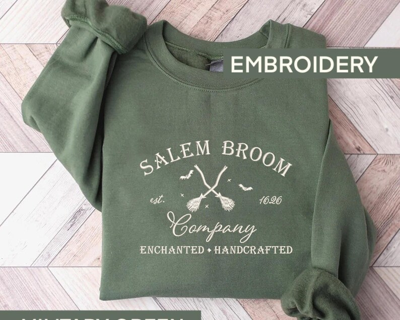 K&ouml;nnte beinhalten: Gr&uuml;ner Pullover mit dem Text "Salem Broom Company est. 1626 Enchanted + Handcrafted" und einer Grafik von zwei gekreuzten Besen mit Flederm&auml;usen, die um sie herum fliegen.