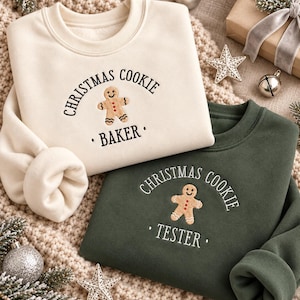 May include: A cream-coloured sweatshirt with black text reading CHRISTMAS COOKIE BAKER and a gingerbread man graphic, paired with a dark green sweatshirt with white text reading CHRISTMAS COOKIE TESTER and a gingerbread man graphic.