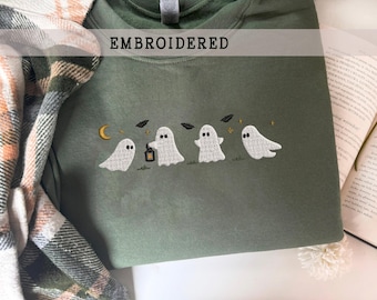 May include: An olive green crewneck sweatshirt featuring an embroidered design of four white ghosts, a yellow crescent moon, small stars, and black bats. The word EMBROIDERED is printed in black text across the upper center of the garment.