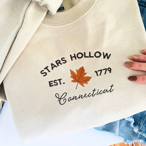 Peut inclure: Un sweat-shirt à col rond de couleur crème avec une broderie marron foncé indiquant Stars Hollow Est. 1779 Connecticut. Une feuille d'érable orange et marron est brodée au centre du texte. Le tissu présente une texture lisse.