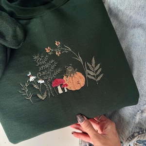 Op de afbeelding: Donkergroene sweatshirt met geborduurd herfstontwerp. De borduursels tonen een pompoen, paddenstoelen en bladeren in oranje, rood en groen. De sweatshirt is gemaakt van een zacht materiaal.