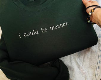 Könnte beinhalten: Ein dunkelwaldgrünes Sweatshirt mit Rundhalsausschnitt und dem weißen Schriftzug "i could be meaner." auf der Brust. Das Kleidungsstück hat lange Ärmel, einen gerippten Halsausschnitt und eine Standardpassform mit schlichtem Design.