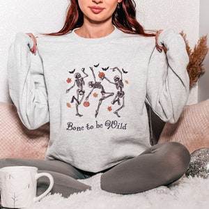 K&ouml;nnte beinhalten: Ein graues Sweatshirt mit einem Motiv von drei tanzenden Skeletten, die K&uuml;rbisse halten. Der Text "Bone to be Wild" ist unter dem Motiv gedruckt.