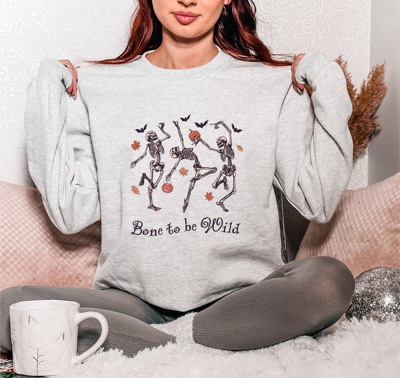 K&ouml;nnte beinhalten: Ein graues Sweatshirt mit einem Motiv von drei tanzenden Skeletten, die K&uuml;rbisse halten. Der Text "Bone to be Wild" ist unter dem Motiv gedruckt.