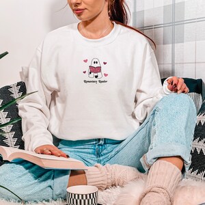 May include: White sweatshirt with a ghost reading a book and pink hearts. The text "Rosemary Reader" is printed below the ghost.