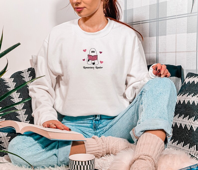 May include: White sweatshirt with a ghost reading a book and pink hearts. The text "Rosemary Reader" is printed below the ghost.