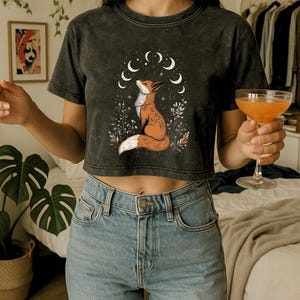 May include: Dark gray crop top featuring a fox illustration with a crescent moon design. The fox is depicted in shades of orange and white, surrounded by floral elements. The shirt is paired with light blue jeans.