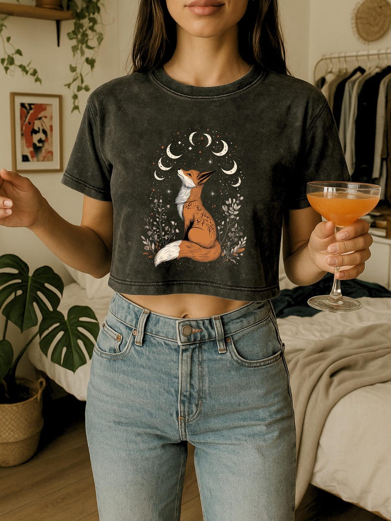 May include: Dark gray crop top featuring a fox illustration with a crescent moon design. The fox is depicted in shades of orange and white, surrounded by floral elements. The shirt is paired with light blue jeans.