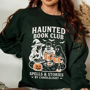 May include: Dark green sweatshirt with a white graphic that reads "HAUNTED BOOK CLUB" above illustrations of a witch, skull, and ghost reading books. The graphic also includes a jack-o'-lantern, bats, and the text "SPELLS & STORIES BY CANDLELIGHT."