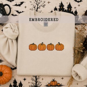 May include: A beige crewneck sweatshirt featuring an embroidered design of four orange pumpkins in a horizontal row across the center. The word EMBROIDERED is printed in black capital letters above the pumpkin graphic on the fabric.