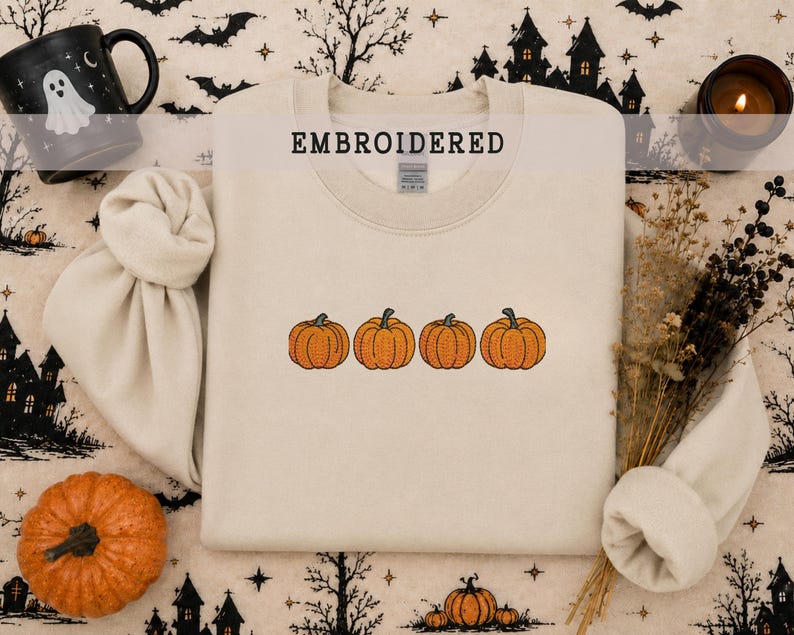 May include: A beige crewneck sweatshirt featuring an embroidered design of four orange pumpkins in a horizontal row across the center. The word EMBROIDERED is printed in black capital letters above the pumpkin graphic on the fabric.