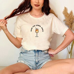 Puede incluir: Una camiseta blanca con el texto "Made for Aperol Spritz" y un gr&aacute;fico de una copa de Aperol Spritz.