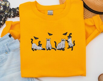 以下が含まれることがあります： 5匹の幽霊猫が魔女の帽子をかぶり、コウモリが飛んでいる刺繍のデザインが施された黄色のスウェットシャツ。スウェットシャツにはギルダンブランドのタグが付いています。