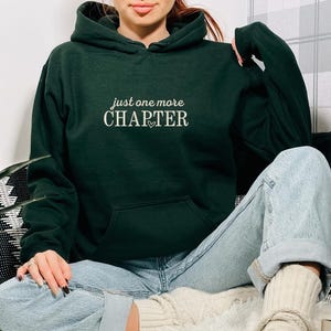 Pode incluir: Um moletom verde escuro com a frase "just one more CHAPTER" em branco, com um pequeno cora&ccedil;&atilde;o. O moletom tem um bolso frontal e um capuz. A pessoa est&aacute; usando jeans azul claro.