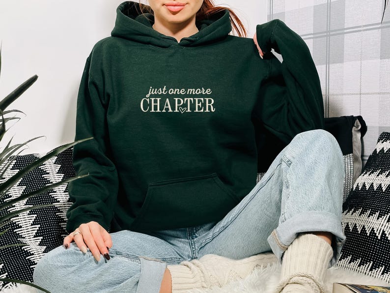 Pode incluir: Um moletom verde escuro com a frase "just one more CHAPTER" em branco, com um pequeno cora&ccedil;&atilde;o. O moletom tem um bolso frontal e um capuz. A pessoa est&aacute; usando jeans azul claro.