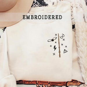 May include: A white sweatshirt with an embroidered design featuring a wand, lightning bolt, glasses, and a snitch. The text "EMBROIDERED" is at the top of the sweatshirt.