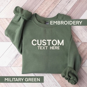 Könnte beinhalten: Ein militärgrünes Langarm-Sweatshirt mit dem gestickten Text CUSTOM TEXT HERE in hellem Cremeweiß. Das Kleidungsstück verfügt über einen Rundhalsausschnitt und gerippte Ärmelbündchen und präsentiert ein klares, schlichtes Design für den Alltag.