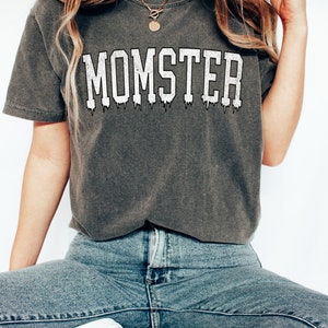 May include: A gray t-shirt with the word "MOMSTER" printed in white with a dripping effect. The shirt is worn by a person with long blonde hair and is paired with blue jeans.