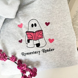 May include: A gray sweatshirt with a white ghost holding a pink book and the text "Romantasy Reader". The ghost is surrounded by pink hearts.
