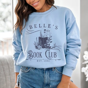 May include: A light blue sweatshirt with a graphic design that says "Belle's Book Club Est. 1991". The design features a book with flowers and vines.