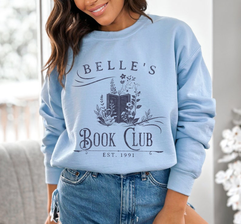 May include: A light blue sweatshirt with a graphic design that says "Belle's Book Club Est. 1991". The design features a book with flowers and vines.