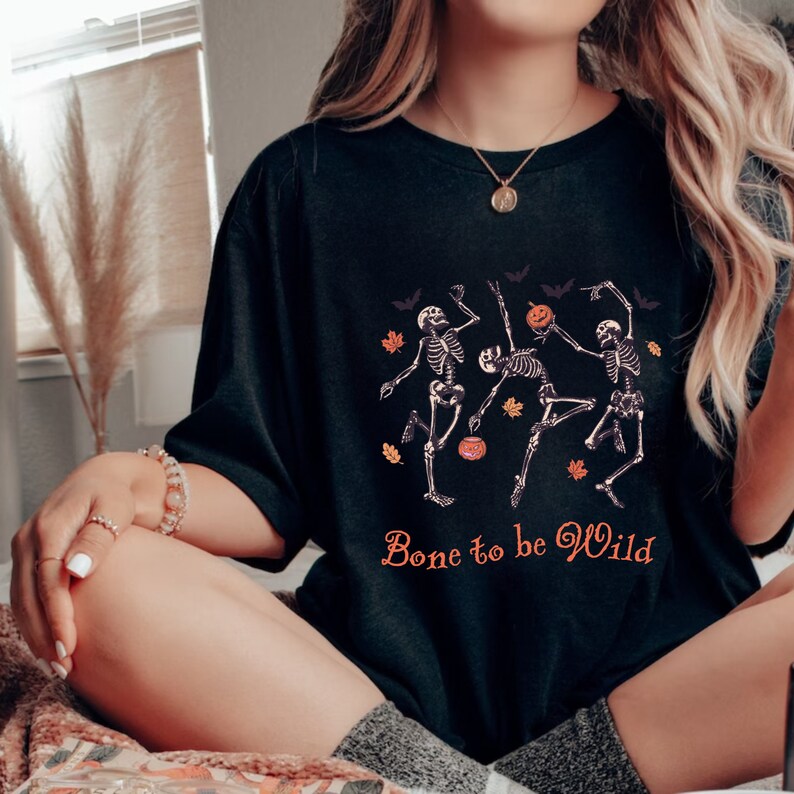 K&ouml;nnte beinhalten: Schwarzes T-Shirt mit einem Motiv von drei Skeletten, die mit K&uuml;rbissen und Flederm&auml;usen tanzen. Der Text "Bone to be Wild" ist unter den Skeletten gedruckt.