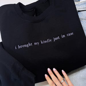 Może przedstawiać: Czarna bluza z okrągłym dekoltem z białym haftowanym napisem "i brought my kindle just in case". Na pierwszym planie widoczna dłoń z pierścionkami. Częściowo widoczne dżinsy. Bluza jest złożona.