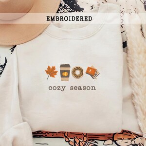 Pode incluir: Camisola creme com gráficos bordados com tema de outono. O design inclui uma folha de bordo, uma chávena de café com uma abóbora, uma rosquinha e livros. As palavras "EMBROIDERED" e "cozy season" também estão na camisola.