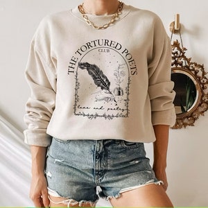 May include: A beige sweatshirt with a black graphic design featuring a quill pen, inkwell, and flowers. The text reads "The Tortured Poets Club" in a black arch.