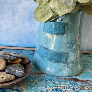 May include: A light blue ceramic vase with a textured surface and white floral designs. The vase has two bands of blue and white patterned trim. A bunch of dried eucalyptus leaves are in the vase.