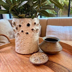 May include: A wooden tray displays a white ceramic vase with dark holes and a textured surface, containing green leafy plants. A small, speckled dish and a bronze-coloured pot are also present.