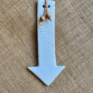 May include: A light blue ceramic arrow with a leather cord and wooden beads. The arrow has a speckled texture and a downward pointing design. The arrow is a decorative item.