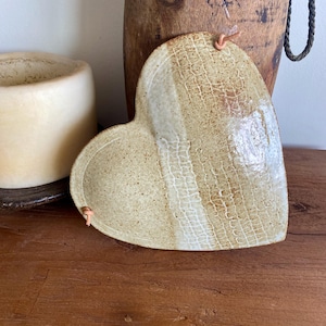 May include: Heart-shaped ceramic dish with a textured surface and a leather loop for hanging. The dish has beige and off-white tones, with a grid-like pattern. A wooden vase and a candle are in the background. The dish is approximately 20 cm wide.