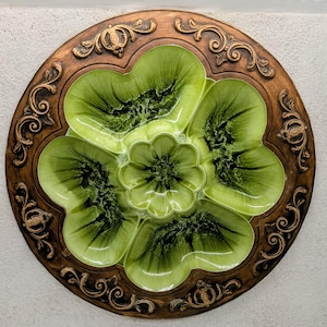 May include: A round, decorative serving dish with a floral design. The dish features a central flower surrounded by five petal-shaped sections in shades of green. The outer rim is a dark brown with ornate, swirling patterns.