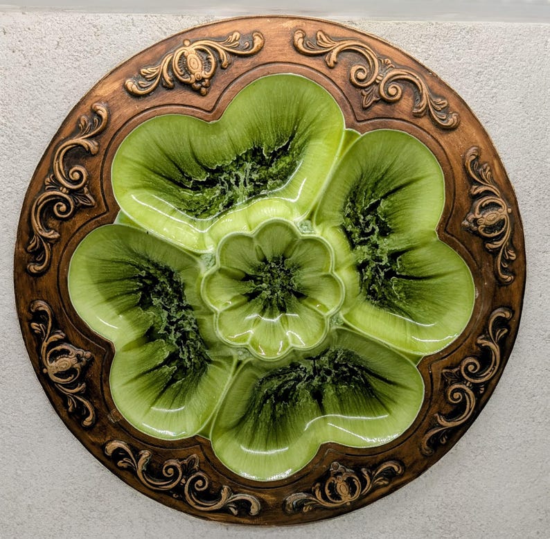 May include: A round, decorative serving dish with a floral design. The dish features a central flower surrounded by five petal-shaped sections in shades of green. The outer rim is a dark brown with ornate, swirling patterns.