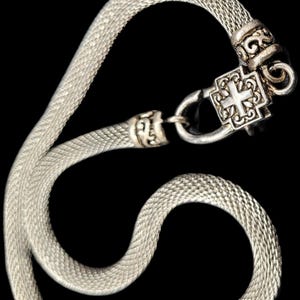 May include: A silver-toned mesh chain necklace featuring a square-shaped clasp with an embossed cross design. The metal chain has a textured, woven appearance and connects to a decorative, ornate metal finding that secures the necklace together.