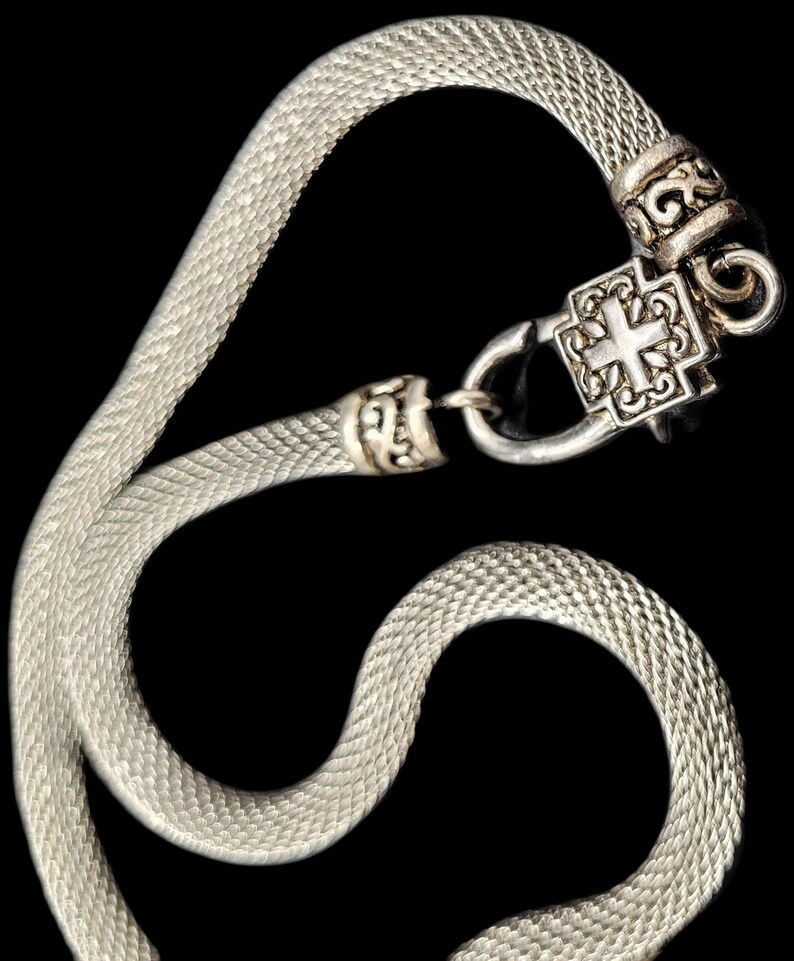 May include: A silver-toned mesh chain necklace featuring a square-shaped clasp with an embossed cross design. The metal chain has a textured, woven appearance and connects to a decorative, ornate metal finding that secures the necklace together.