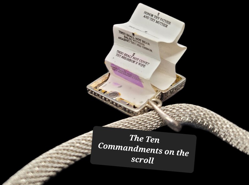 May include: A silver-toned metal locket pendant attached to a textured mesh chain. The locket is open, revealing a white accordion-folded paper scroll printed with black text, including the phrases Honor Thy Father and Thy Mother.