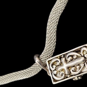 May include: A silver-toned mesh chain necklace featuring a rectangular pendant. The pendant displays an embossed cross motif surrounded by intricate scrollwork patterns. The metal surface has a polished finish with darkened recessed areas.