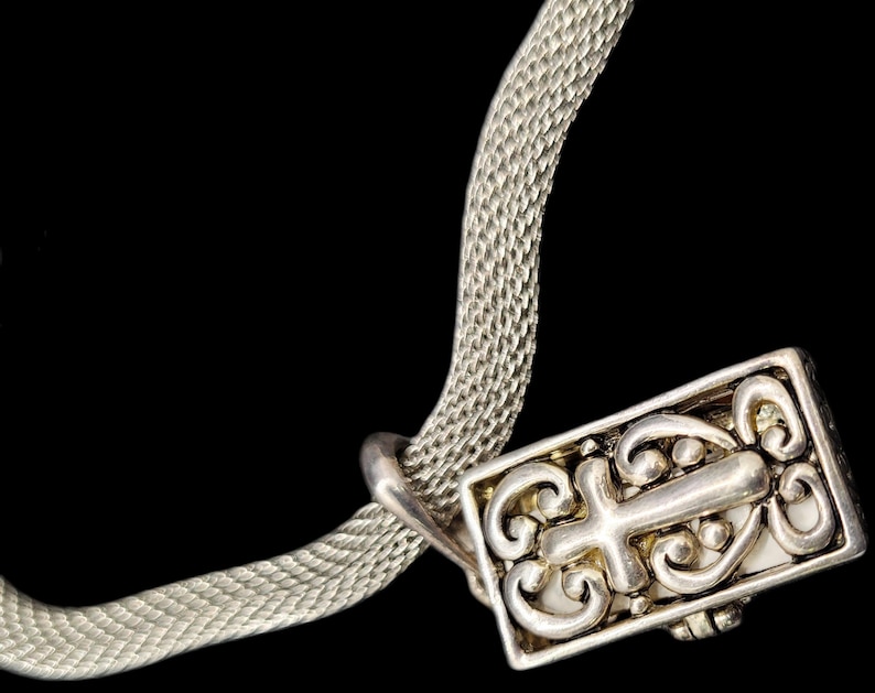 May include: A silver-toned mesh chain necklace featuring a rectangular pendant. The pendant displays an embossed cross motif surrounded by intricate scrollwork patterns. The metal surface has a polished finish with darkened recessed areas.