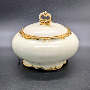 May include: A cream-colored, lidded ceramic jar with gold trim. The jar has a rounded shape with a decorative gold handle on the lid. The gold trim accents the rim and base of the jar, adding a touch of elegance.