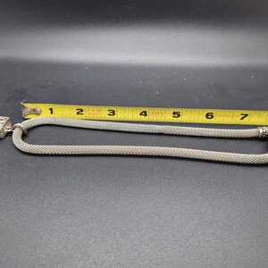 May include: A silver-toned mesh chain necklace featuring a rectangular pendant with an intricate scrollwork pattern. A yellow measuring tape is positioned alongside the necklace, indicating a total length of approximately 8 inches for the chain.